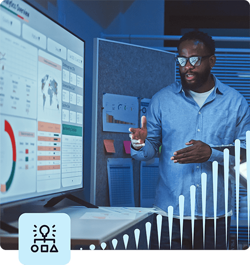 A man wearing glasses stands in a dark room as a presentation is being displayed on a whiteboard. He is speaking with his hands as he gestures towards something on the screen. The screen displays examples of pie charts, maps and tables.